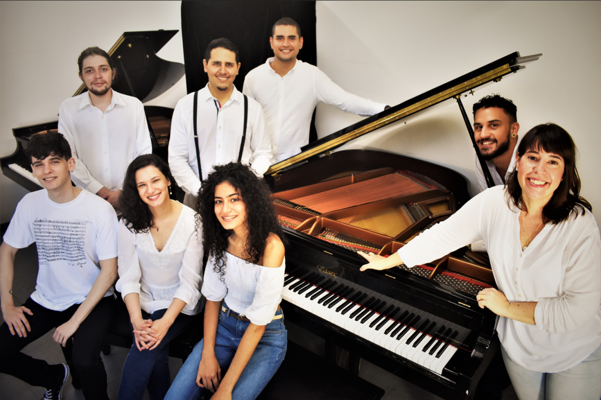 Grupo de pianistas jovens, todos vestindo branco, se reúne ao redor piano. Há 3 mulheres e 5 homens.  